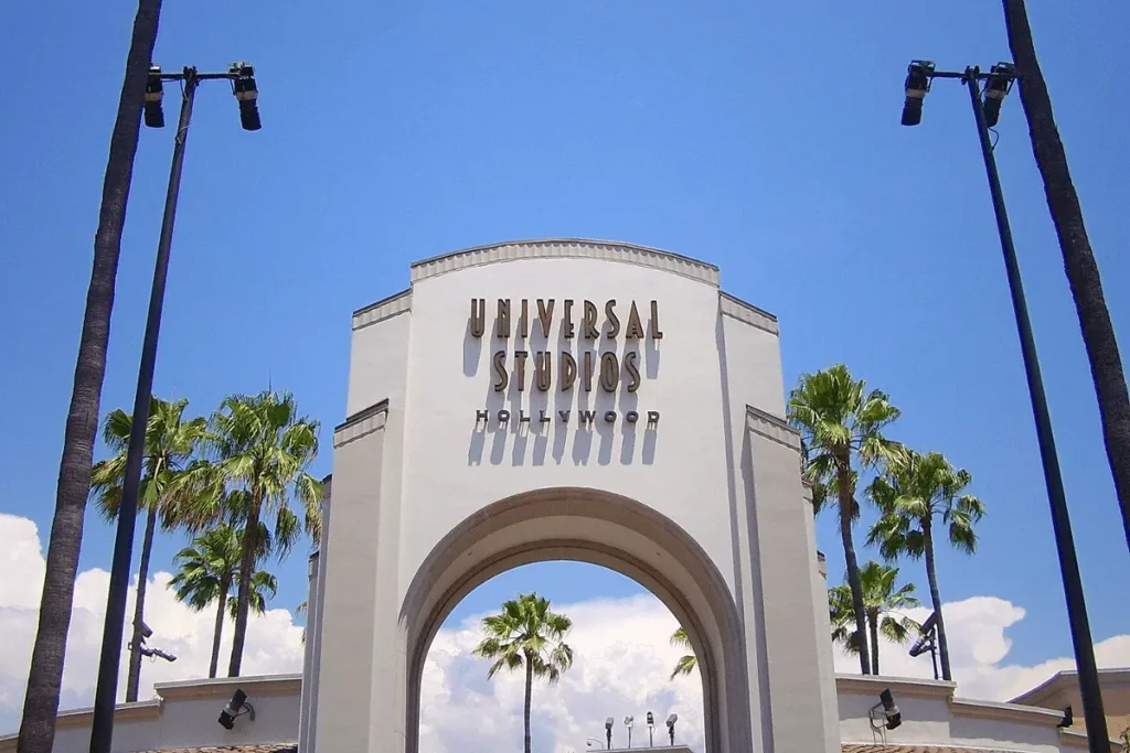 Entrance of Universal Studios Hollywood showing the large sign above the entryway, a paved walkway leading inside, a stone wall on one side, and tall palm trees visible behind the entrance under a clear sky.