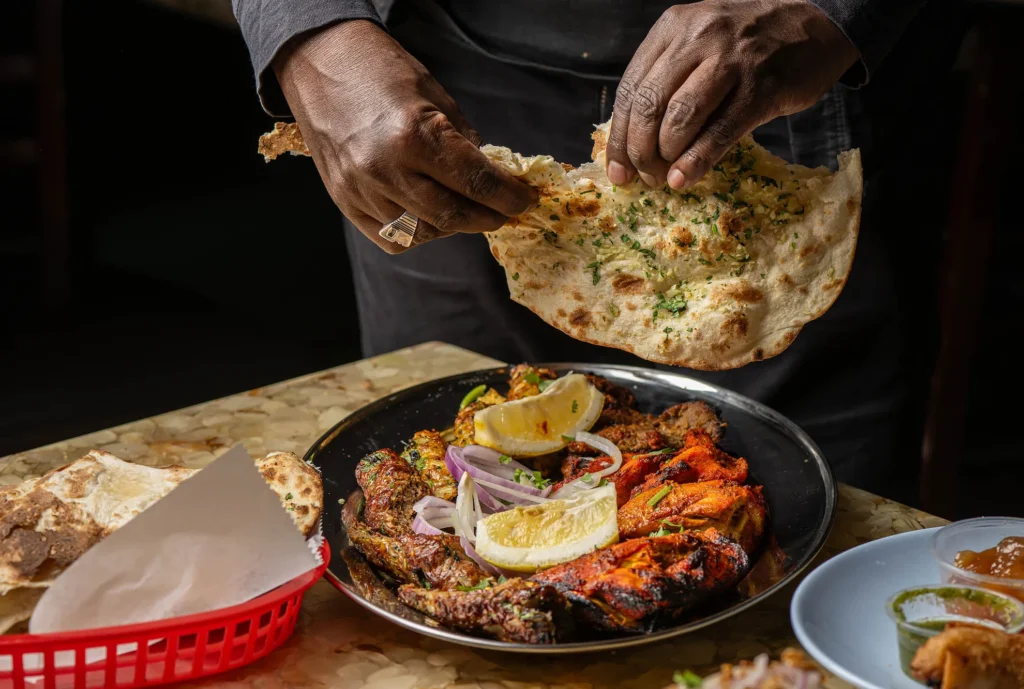 Close-up view of Biriyani Kabob House dishes on a table, including biryani, grilled meats, and sauces, with a hand in the background splitting fresh bread above the meal.