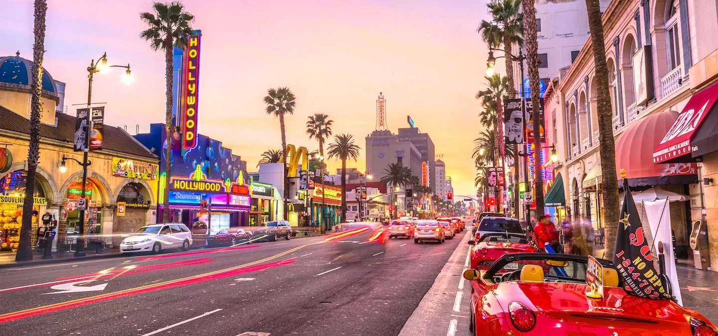 Sunset view of the roads and streets along Hollywood Boulevard, with warm light reflecting off buildings, palm trees lining the sidewalks, and a few cars and pedestrians in motion.