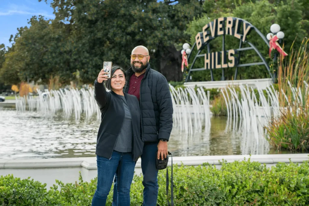 A couple taking a selfie with the iconic Beverly Hills sign in the background, surrounded by greenery and a sunny outdoor setting.