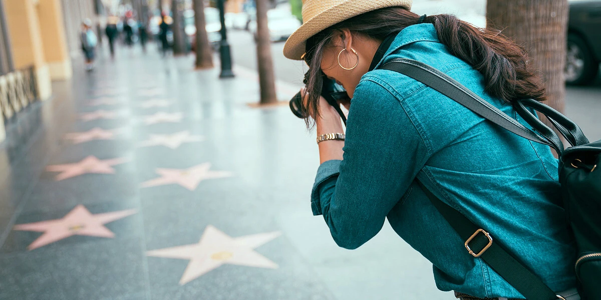 A tourist woman taking a photo of the Hollywood Walk of Fame sidewalk, capturing the stars embedded in the pavement along the bustling street.