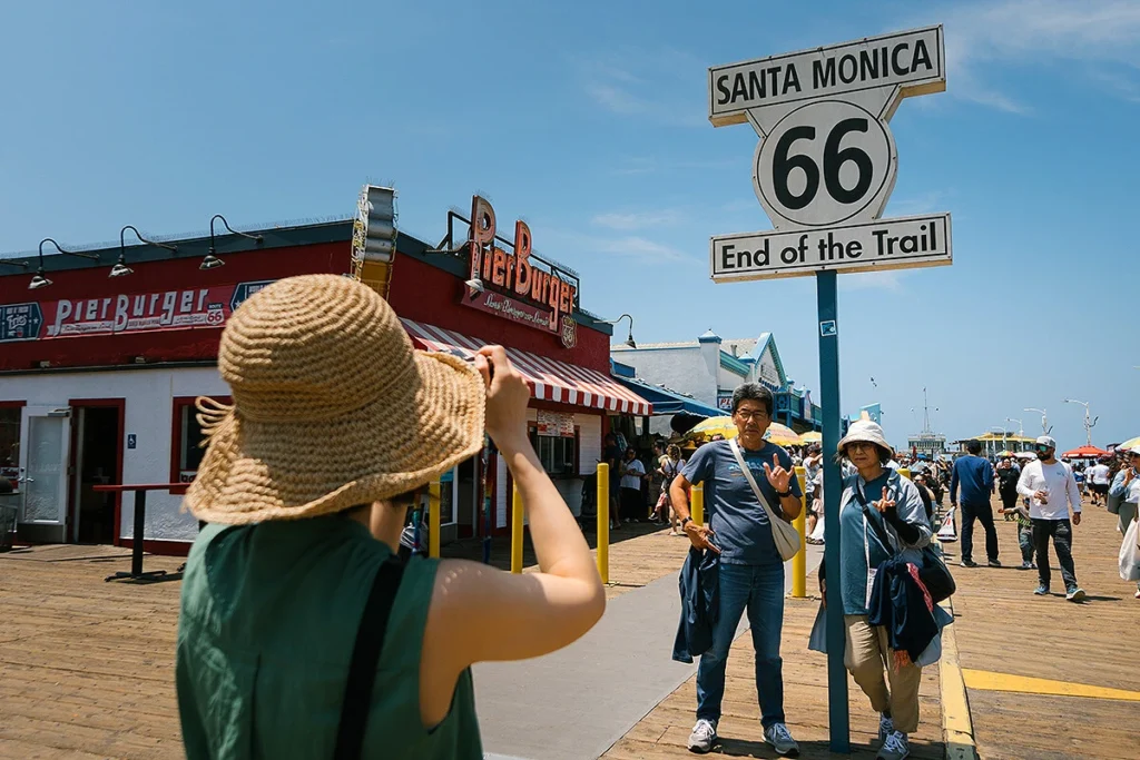 A daughter taking a photo of her smiling parents standing near the historic Santa Monica Route 66 sign, with a clear sky and beachside atmosphere in the background.