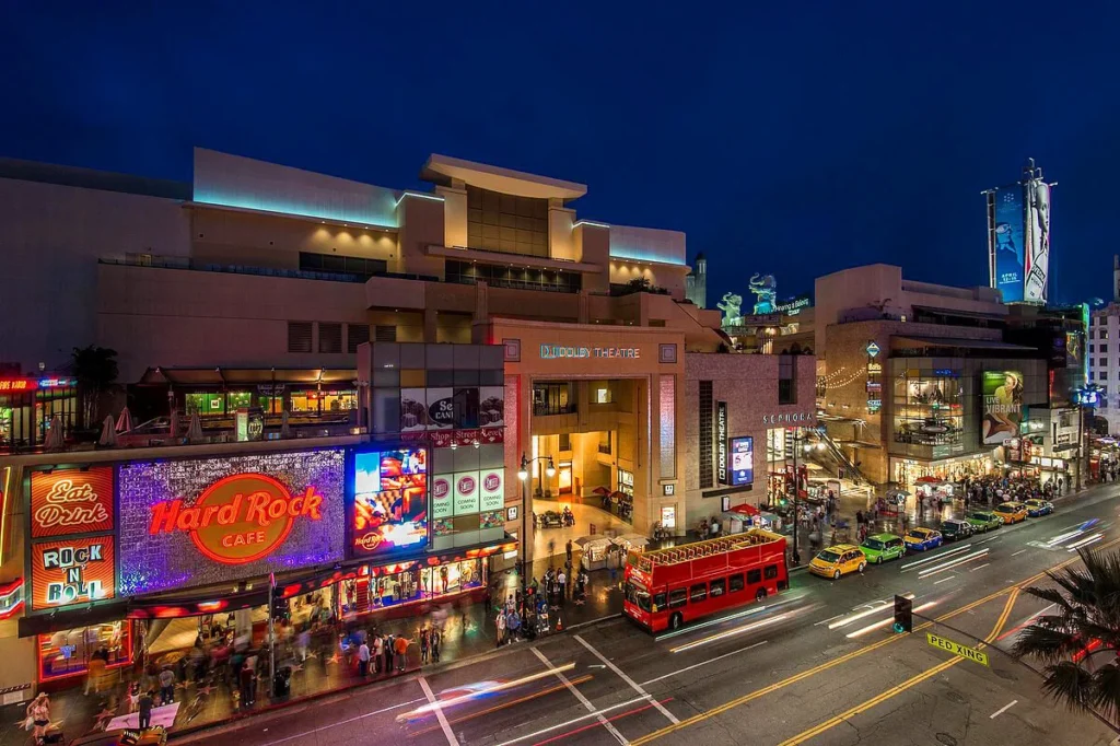 Nighttime exterior view of the Hollywood & Highland Center, brightly lit with decorative lights, showcasing its grand architecture, archways, and the illuminated entrance with bustling crowds.