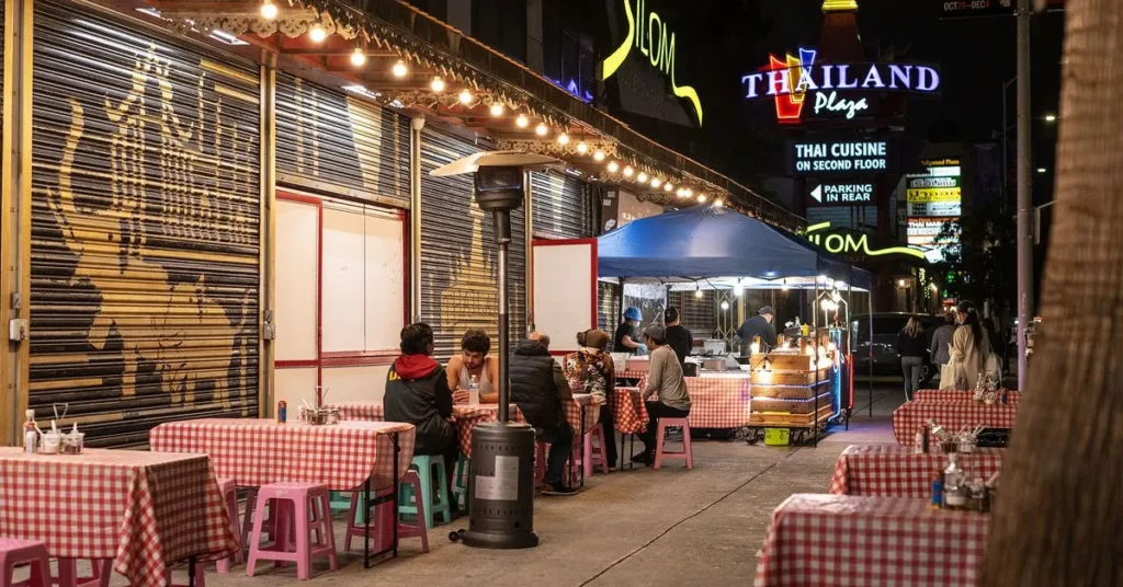 Nighttime view of a street in Thai Town, lined with brightly lit restaurants and food vendors, showcasing colorful signs, busy sidewalks, and a vibrant dining atmosphere.