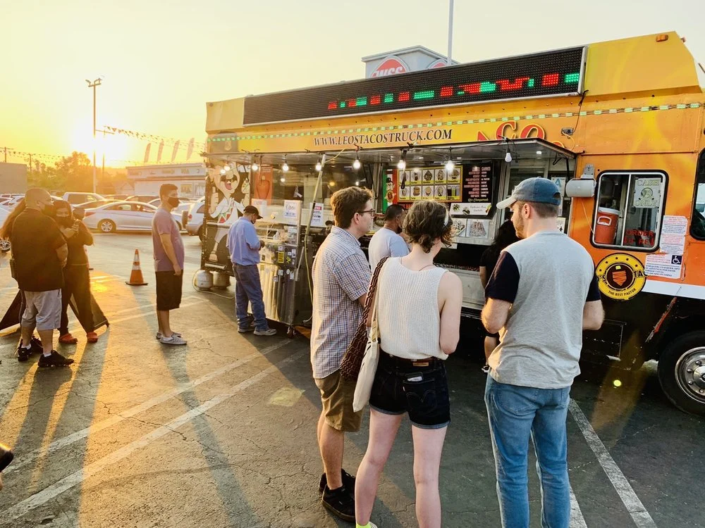 A group of people gathered in front of Leo's Taco Truck in Hollywood during sunset, with warm lighting casting a glow on the crowd and the colorful food truck.