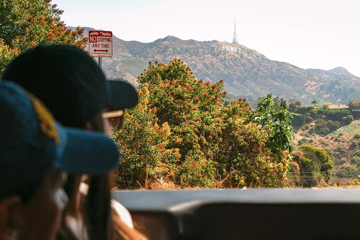 Tourists in a vehicle looking toward the Hollywood Sign across the hills on a sunny day.