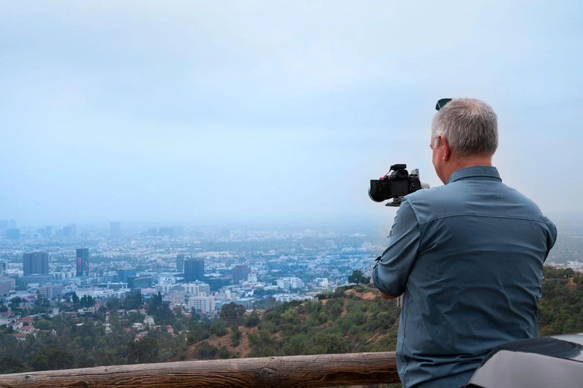 Man with a camera on a tripod at a wooden-railed overlook, capturing a hazy Los Angeles skyline and rolling hills below.