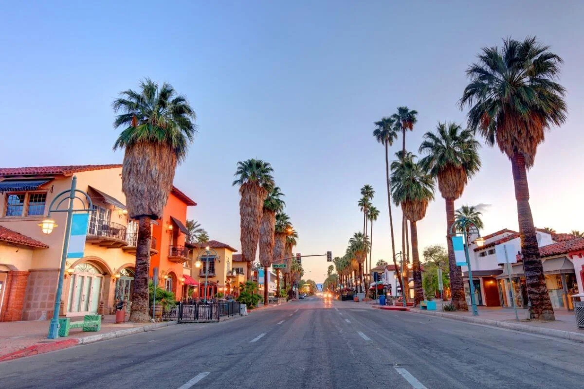 Empty main street in downtown Palm Springs lined with tall palm trees and Spanish-style buildings, glowing in warm sunset light under a clear sky.