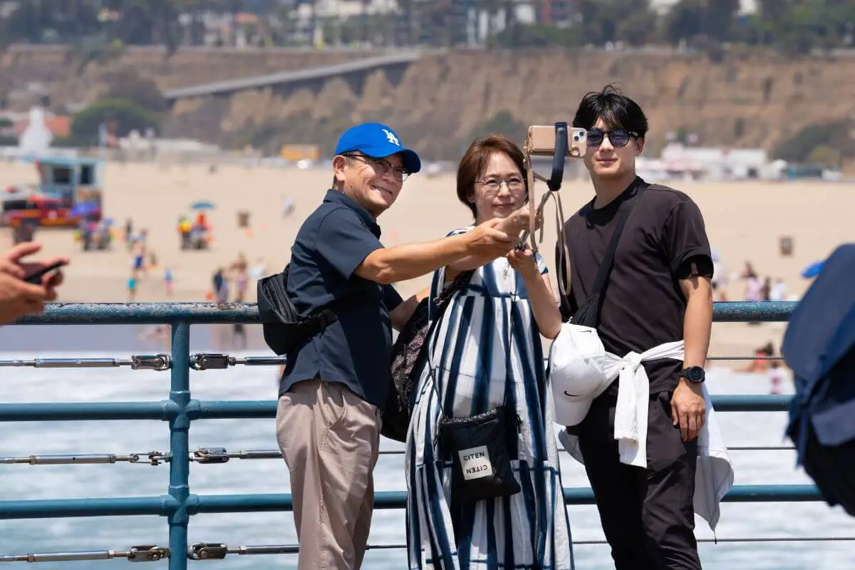 A family taking a selfie with a smartphone on Santa Monica Pier, smiling with the beach and cliffs of Los Angeles in the background on a bright, sunny day.