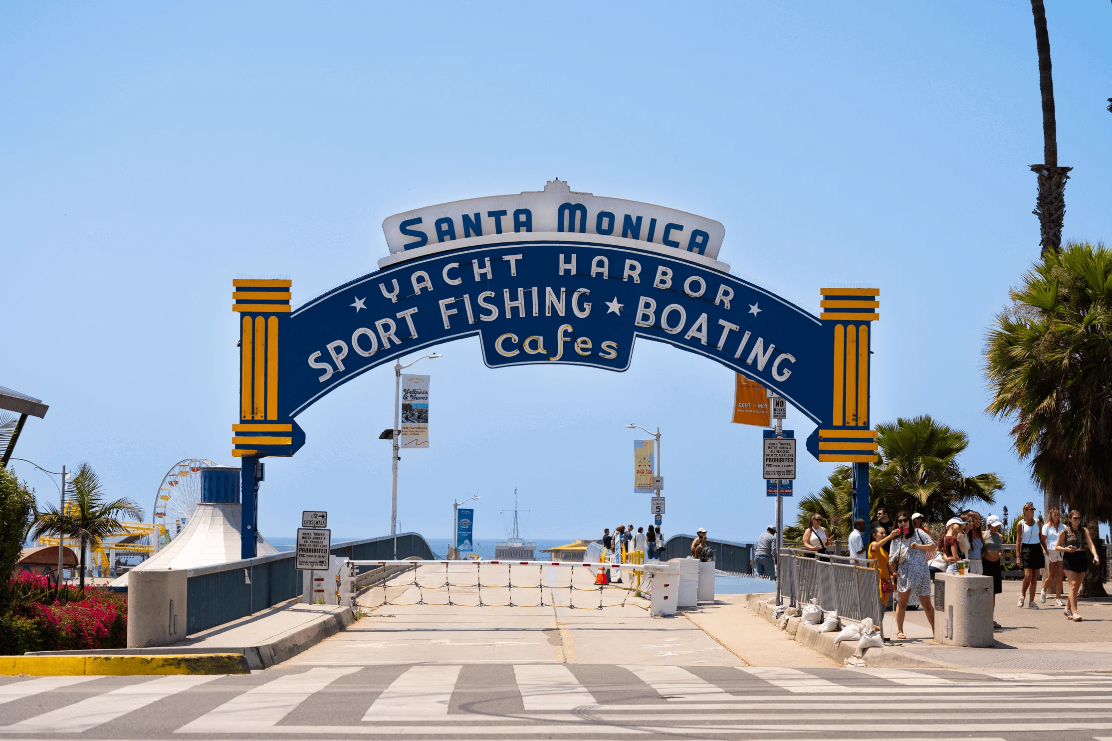 Iconic blue arch reading “Santa Monica Yacht Harbor — Sport Fishing • Boating • Cafes” at the pier entrance, with pedestrians and Pacific Park rides visible under a clear sky.
