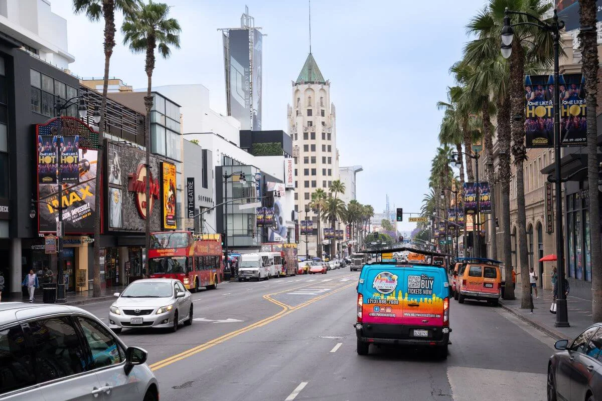 Daytime view along palm-lined Hollywood Boulevard with tour buses, cars, and theater marquees, looking toward a historic tower in the distance.