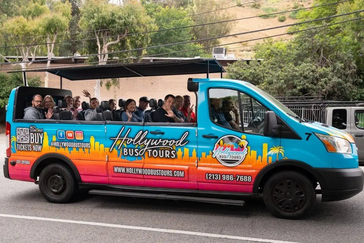 A colorful open-roof Hollywood Bus Tours van driving through Los Angeles with smiling tourists waving, surrounded by trees and hills on a sunny day.