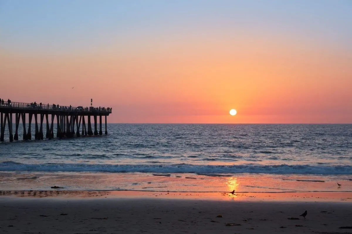 A vibrant sunset over the Pacific Ocean viewed from Hermosa Beach, with the pier extending into the water and silhouettes of people watching the sun dip below the horizon.