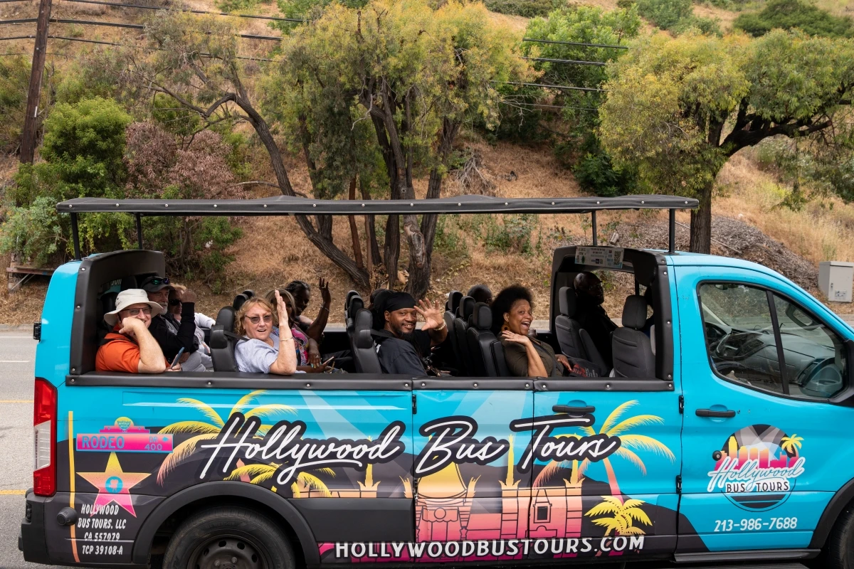 Bright turquoise Hollywood Bus Tours open-air tour vehicle with happy passengers waving, driving through Los Angeles hills on celebrity homes sightseeing tour, featuring tropical palm tree graphics and company branding.