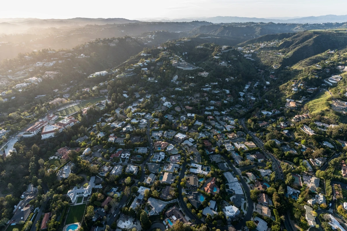 Sweeping aerial panorama of Hollywood Hills at sunset showing densely packed luxury homes nestled in tree-covered hillsides with winding roads, swimming pools, and expansive views across Los Angeles during golden hour.