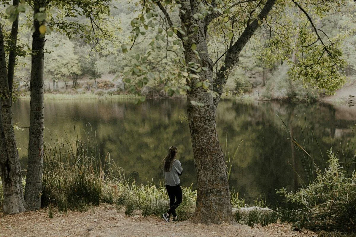 Person standing under leafy trees at the edge of a calm forest lake, looking across reflective water on an overcast day.