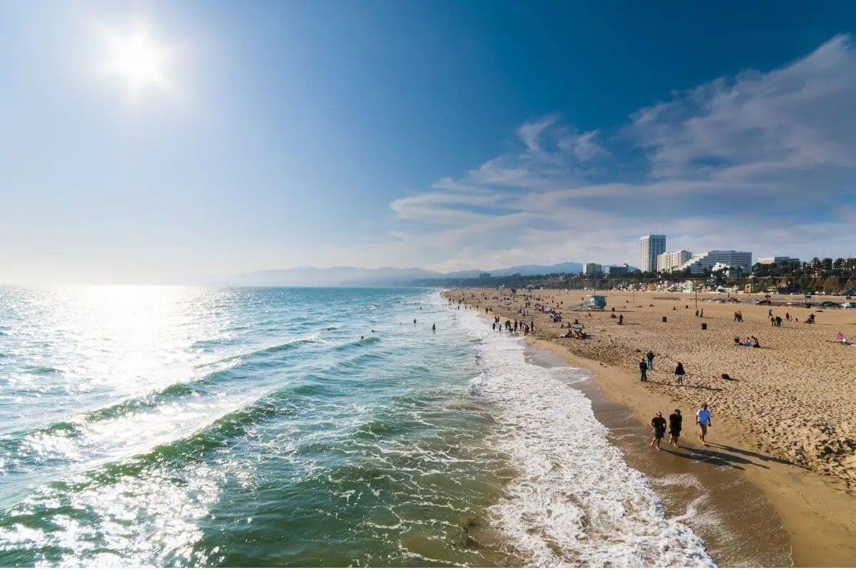 View of Santa Monica Beach on a bright sunny day with people walking along the shoreline, gentle ocean waves, and city buildings visible in the distance under a clear blue sky.