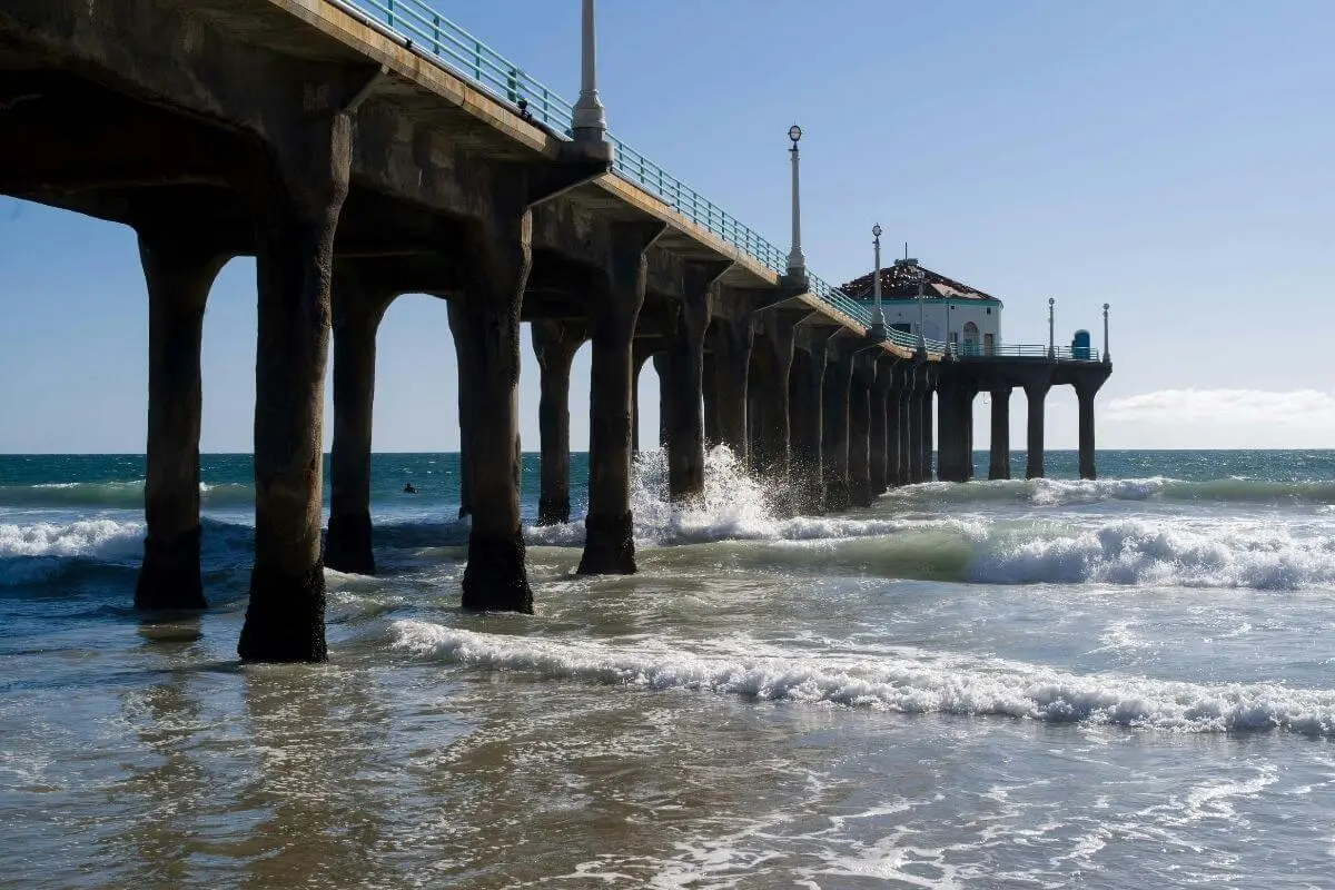View of Manhattan Beach Pier extending over the Pacific Ocean with waves crashing against the pier’s concrete pillars on a bright, sunny day.