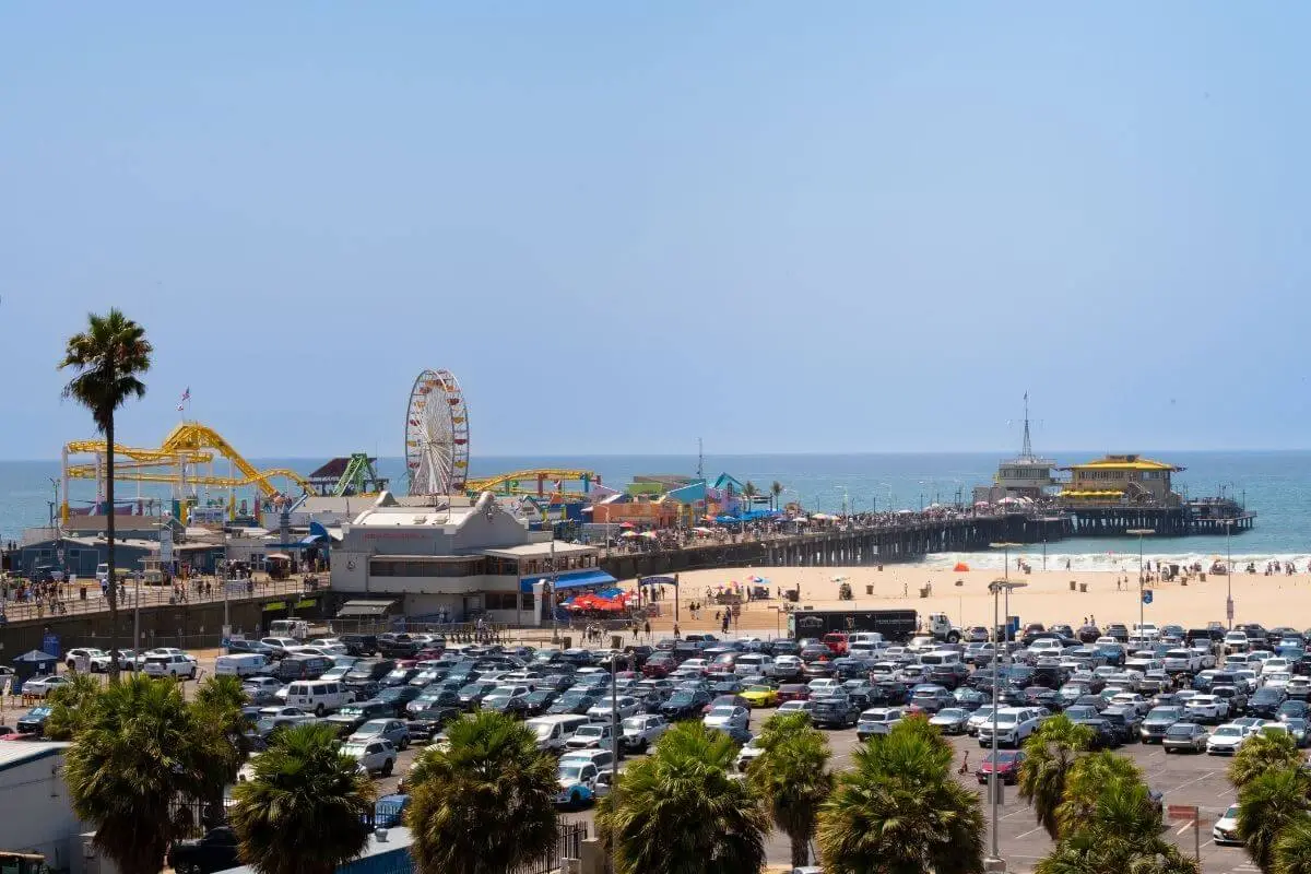 A bright view of Santa Monica Pier in California featuring the iconic Ferris wheel, roller coaster, and beachgoers enjoying the sand and ocean under a clear blue sky, with a parking lot filled with cars in the foreground.