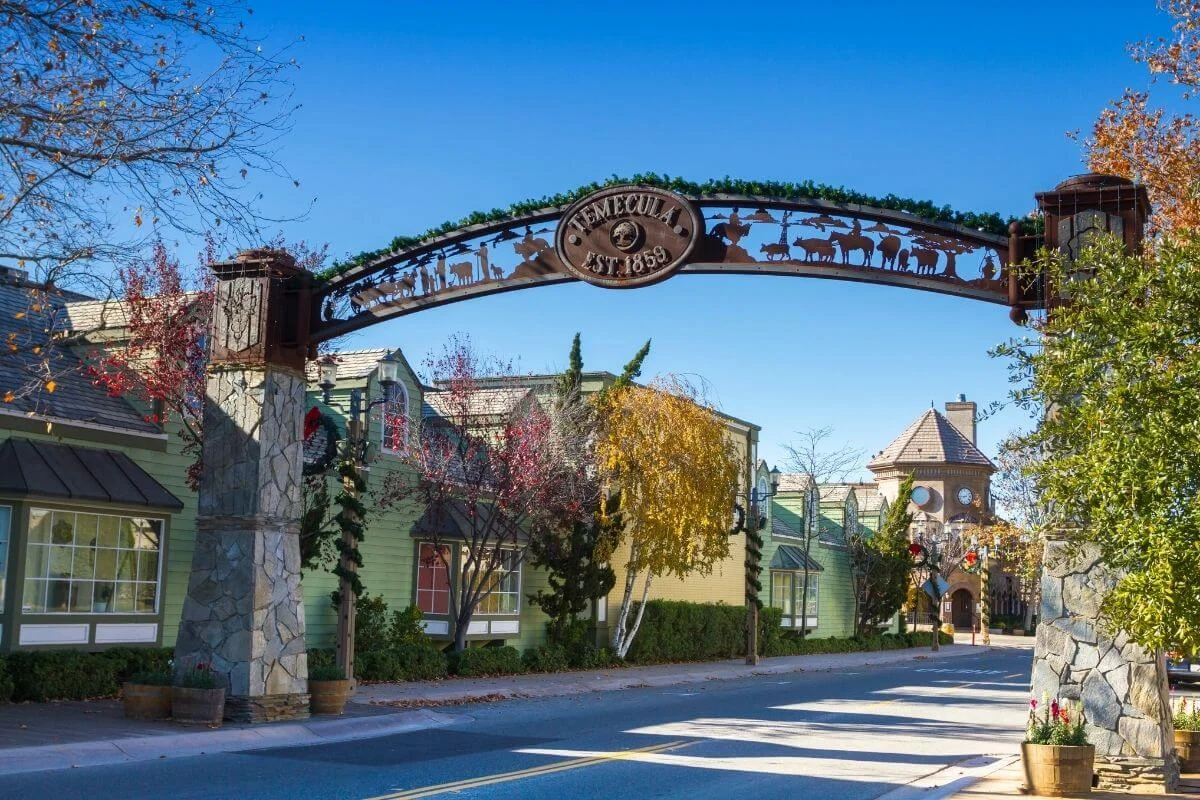 Decorative metal arch reading “Temecula Est. 1859” spans a quiet street lined with quaint storefronts and autumn trees in Old Town Temecula under a bright blue sky.