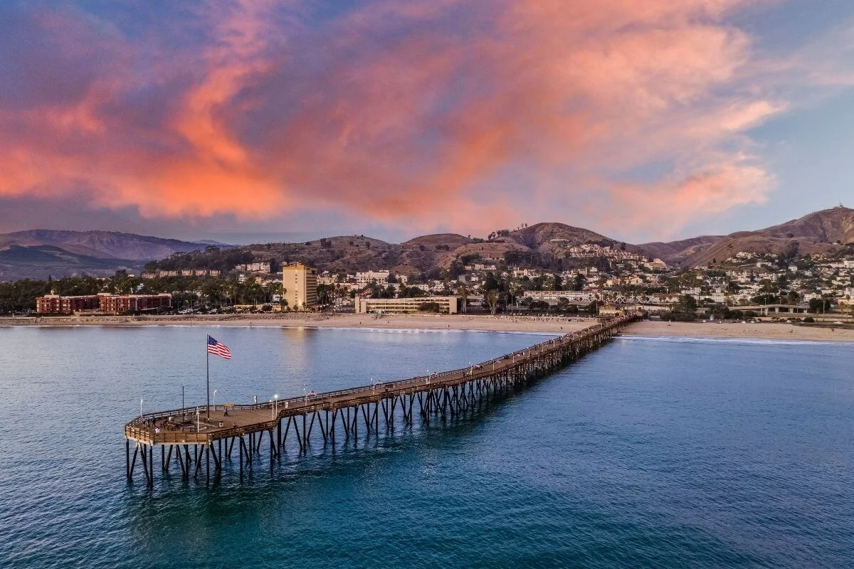 A long wooden pier with an American flag stretches into calm ocean waters at sunset, with a coastal town and rolling hills under pink clouds in the background.