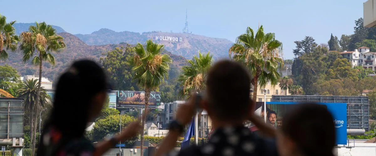 Blurred tourists take photos of the distant Hollywood Sign framed by palm trees on a sunny day in Los Angeles, California.
