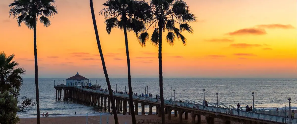 Palm trees framing Manhattan Beach Pier during a colorful sunset over the Pacific Ocean, with people walking along the pier and relaxing on the beach.