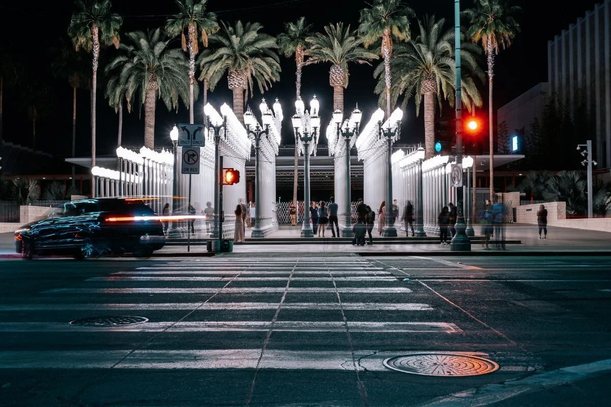 Long exposure night photo of people walking among rows of glowing white street lamps at the Urban Light installation, with palm trees behind and cars and a crosswalk in the foreground.