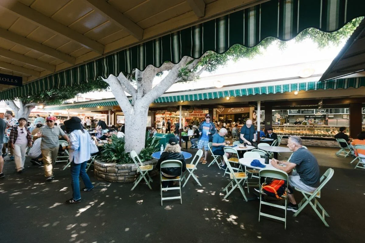People sit and walk through a shaded outdoor food court at a farmers market, with green-and-white striped awnings, white trees in planters, and folding tables and chairs scattered around.