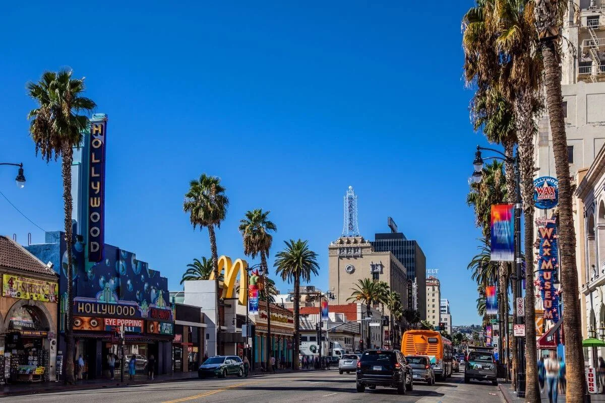 Palm-lined Hollywood Boulevard with cars, neon signs, and the Hollywood Wax Museum under a bright blue sky.