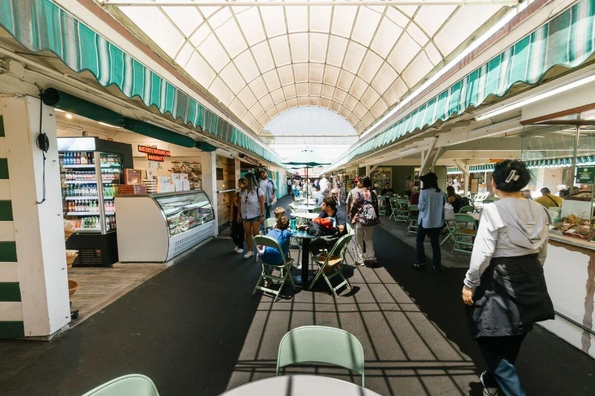 People walk and sit at tables under a bright arched canopy between two rows of farmers market food stalls, with green-striped awnings, display cases, and a small grocery counter on the left.