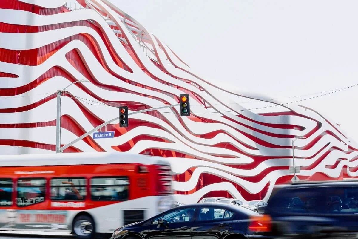 Street view of cars and a red city bus moving along Wilshire Boulevard in front of the Petersen Automotive Museum, showing its bold red and white wavy metal exterior.