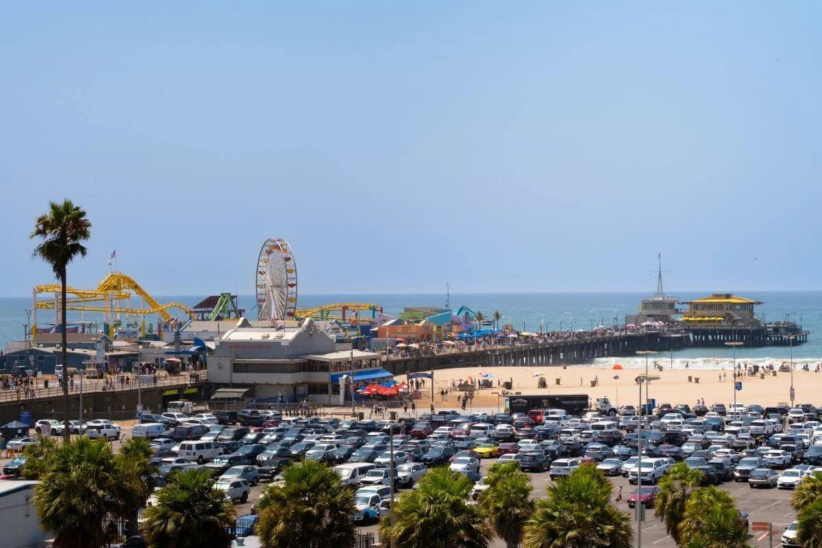 Wide view of Santa Monica Pier with the Pacific Park Ferris wheel, busy beach, and full parking lot under a clear blue sky.