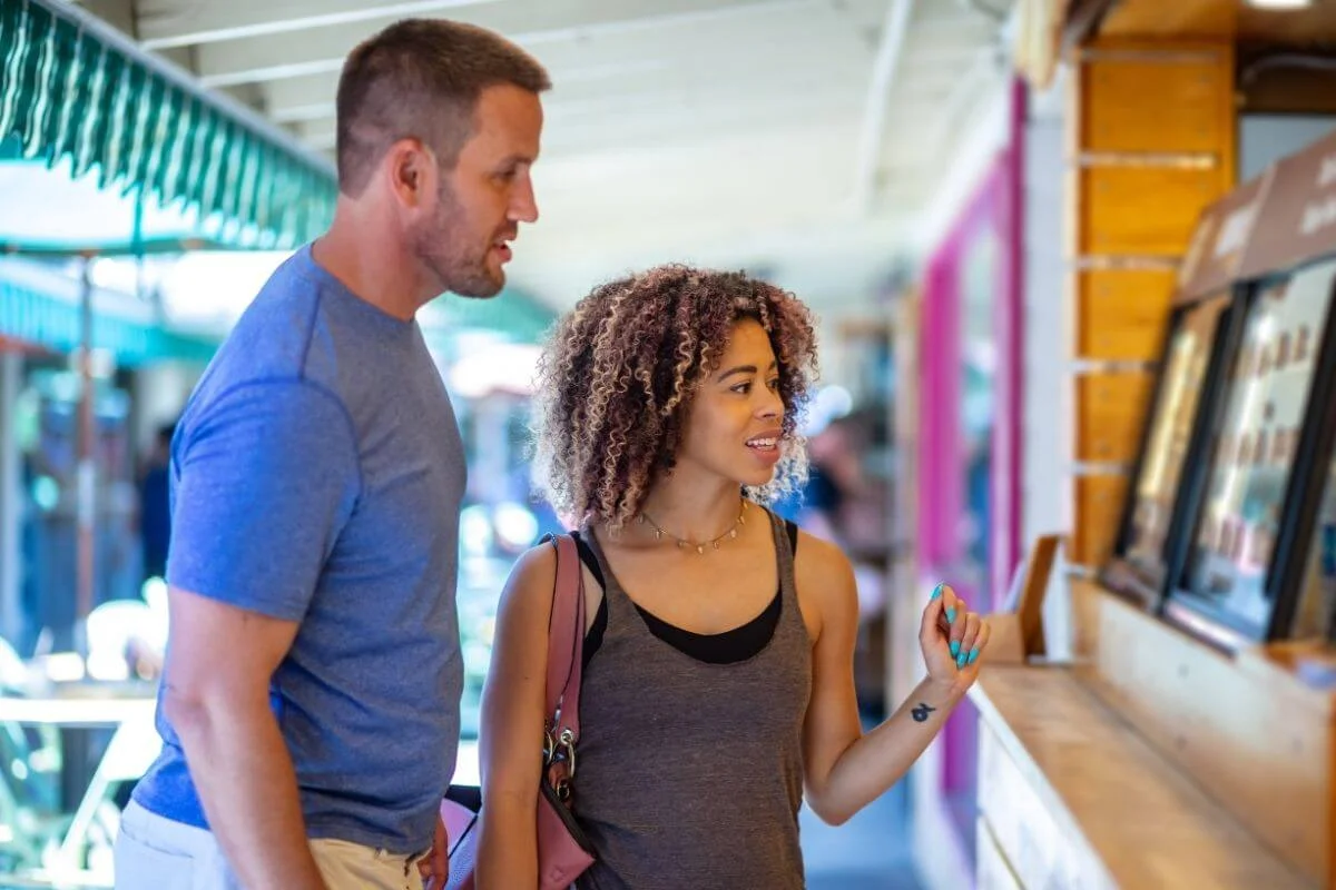 Man and woman stand side by side at an outdoor market counter, looking at a menu display as the woman gestures while deciding what to order.