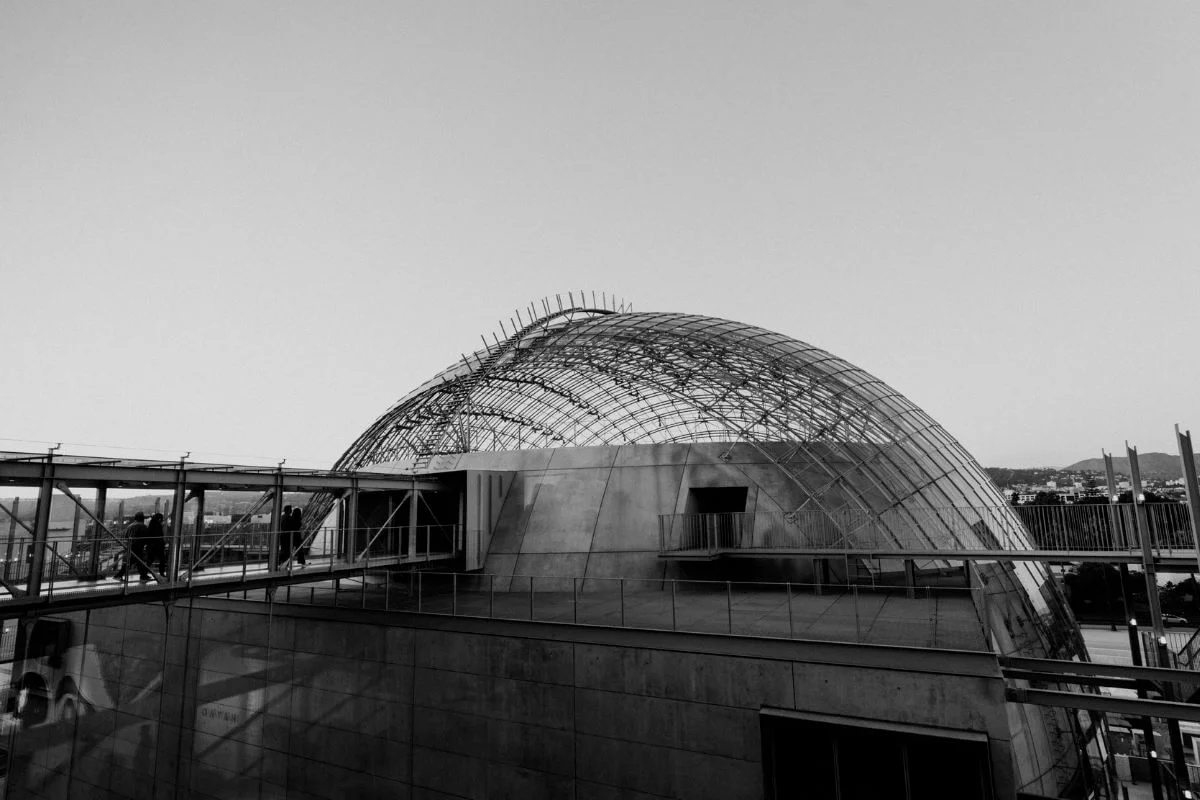 Black and white photo of a modern concrete building with a large metal lattice dome on top, connected by elevated walkways where small figures are walking, against a clear open sky.