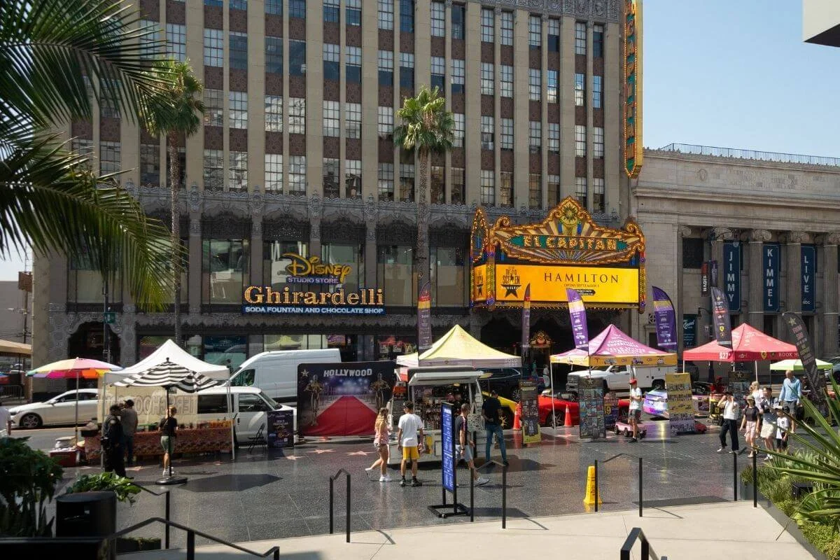 Daytime view of Hollywood Boulevard with the El Capitan Theatre marquee, Disney Ghirardelli store, vendor tents, and crowds along the Hollywood Walk of Fame. 
