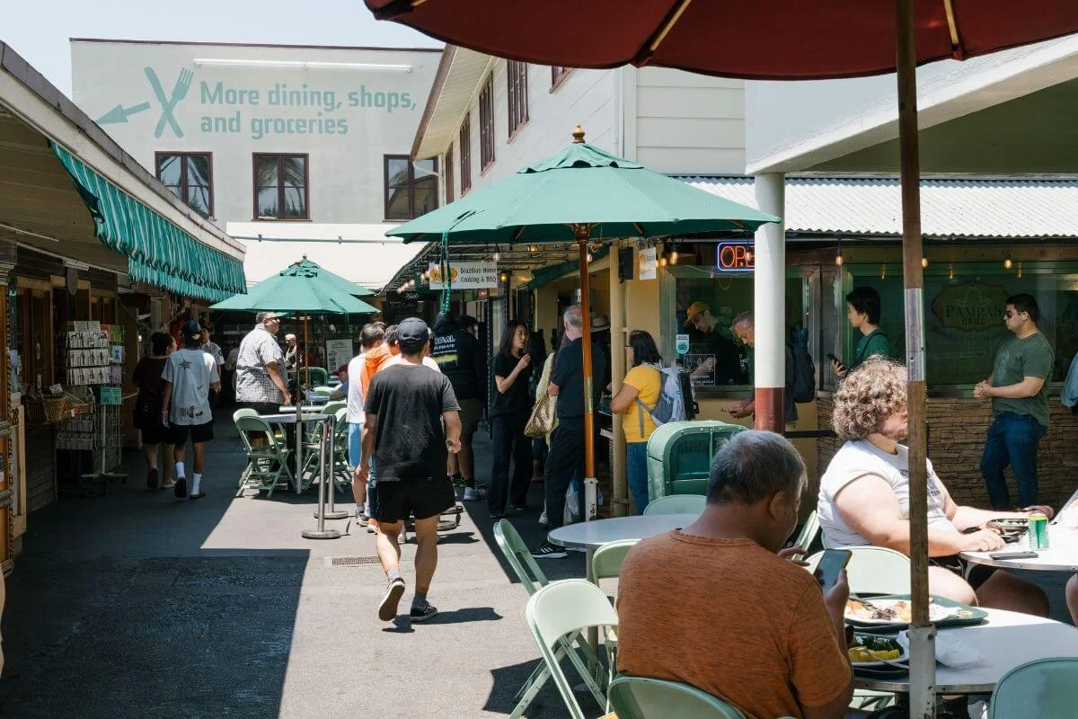People walk and dine in a narrow outdoor market alley lined with food stalls, green umbrellas, and folding tables, with a large wall sign in the background reading “More dining, shops, and groceries.”