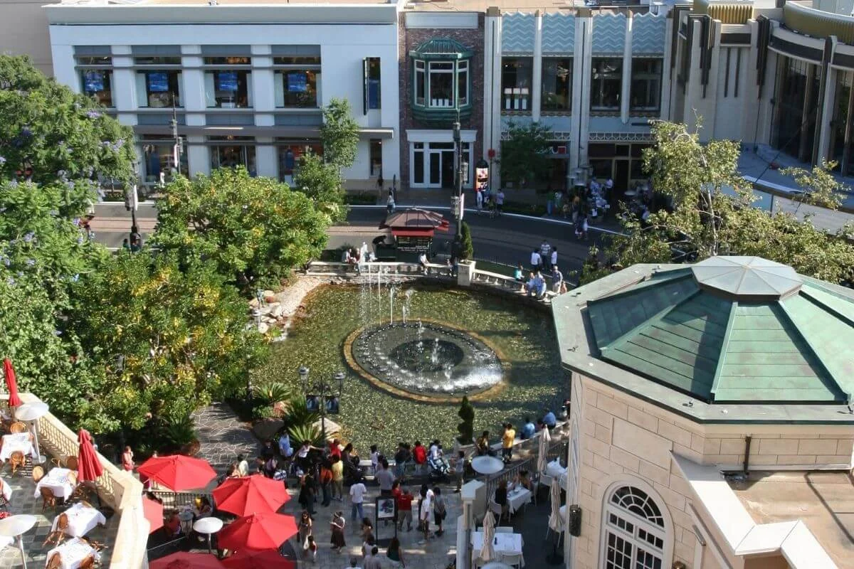 Aerial view of a round fountain surrounded by trees, crowds of people, and outdoor café tables with red umbrellas, set in the middle of a stylish open-air shopping district.
