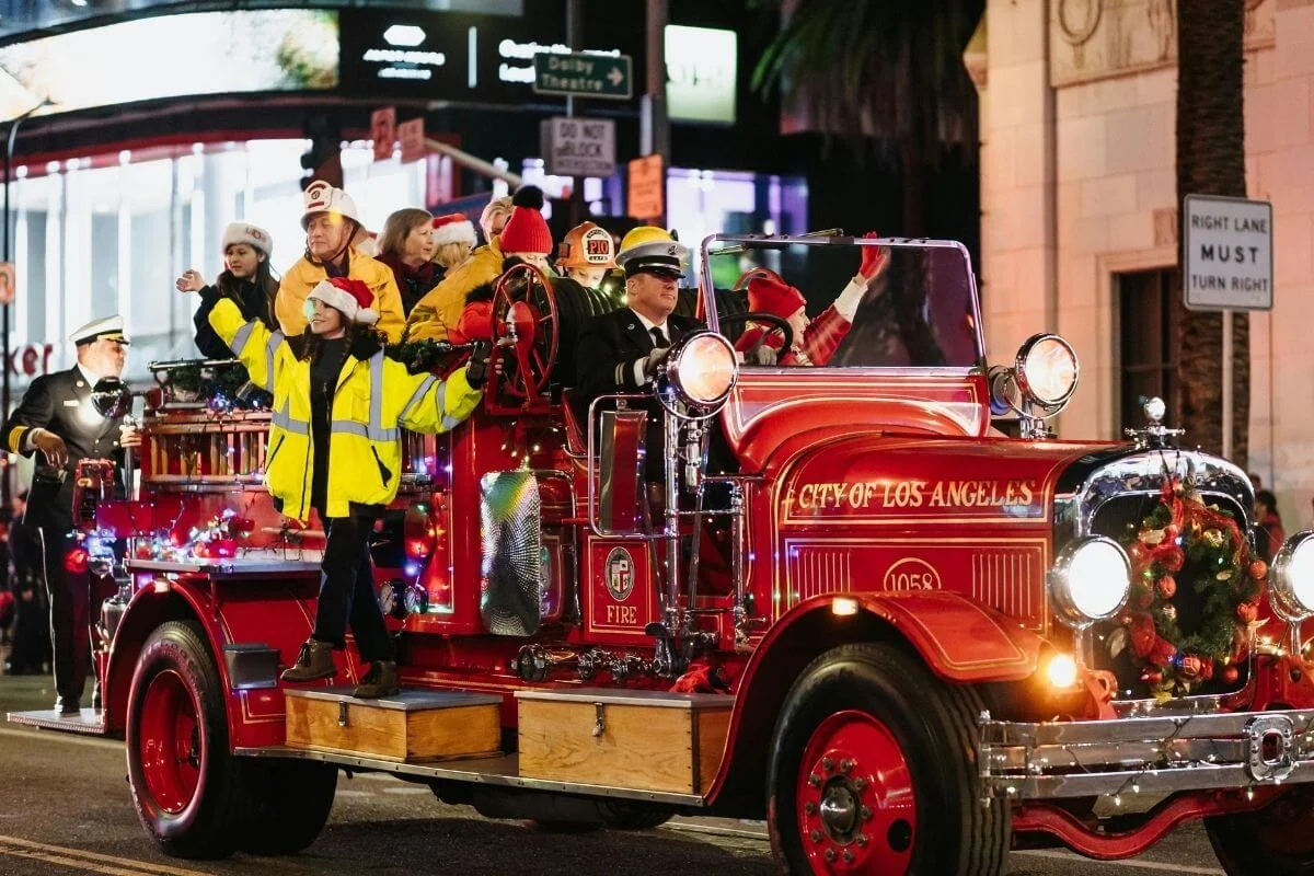 Vintage red Los Angeles fire truck decorated with lights and wreaths carries smiling participants in Santa hats during a nighttime holiday parade.