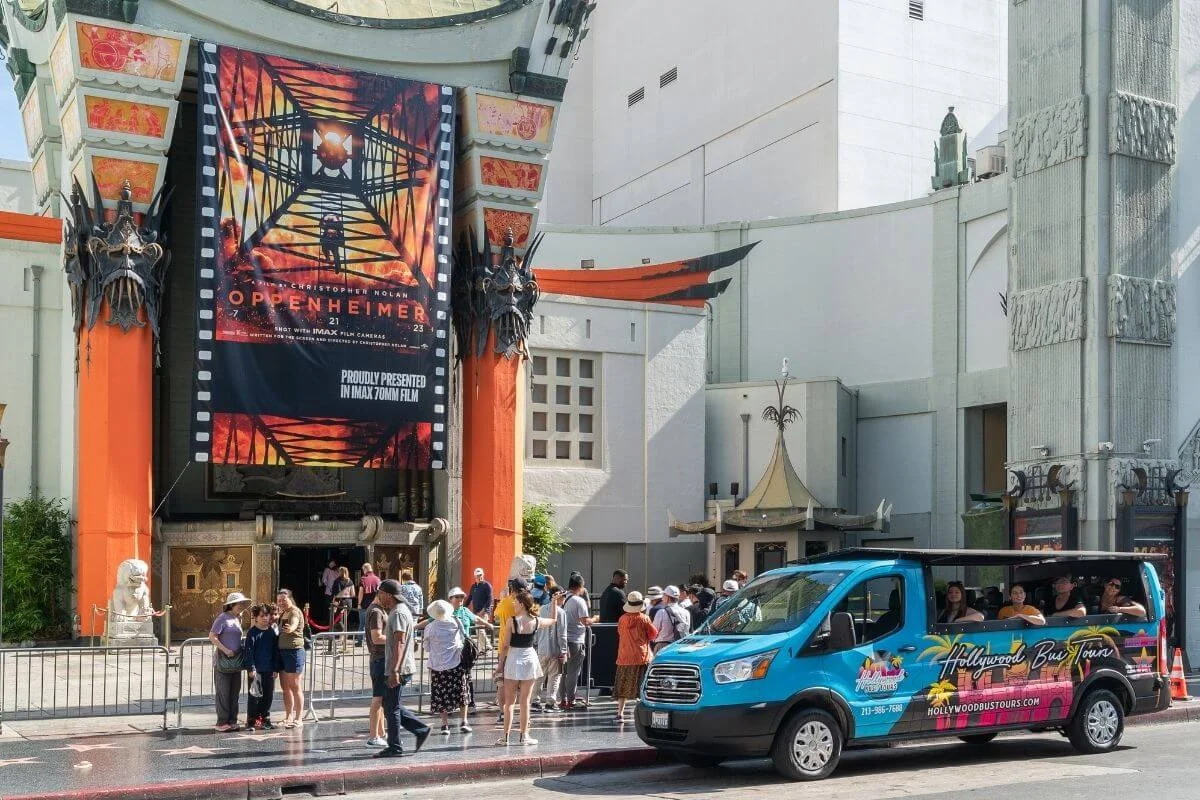 Blue open-air tour van drives past the TCL Chinese Theatre on Hollywood Boulevard, with crowds and a large movie poster visible at the entrance.