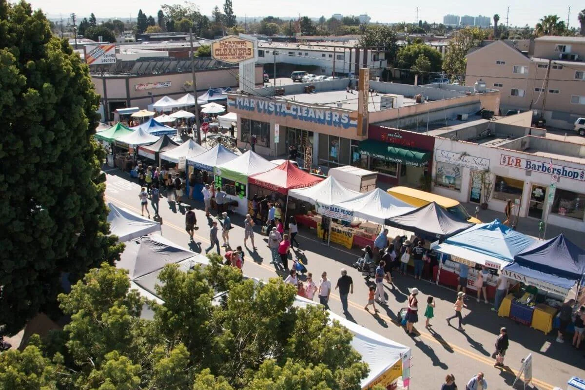Aerial view of a busy street market lined with rows of colorful canopy tents in front of small shops, as people walk between stalls and browse vendors on a sunny day.