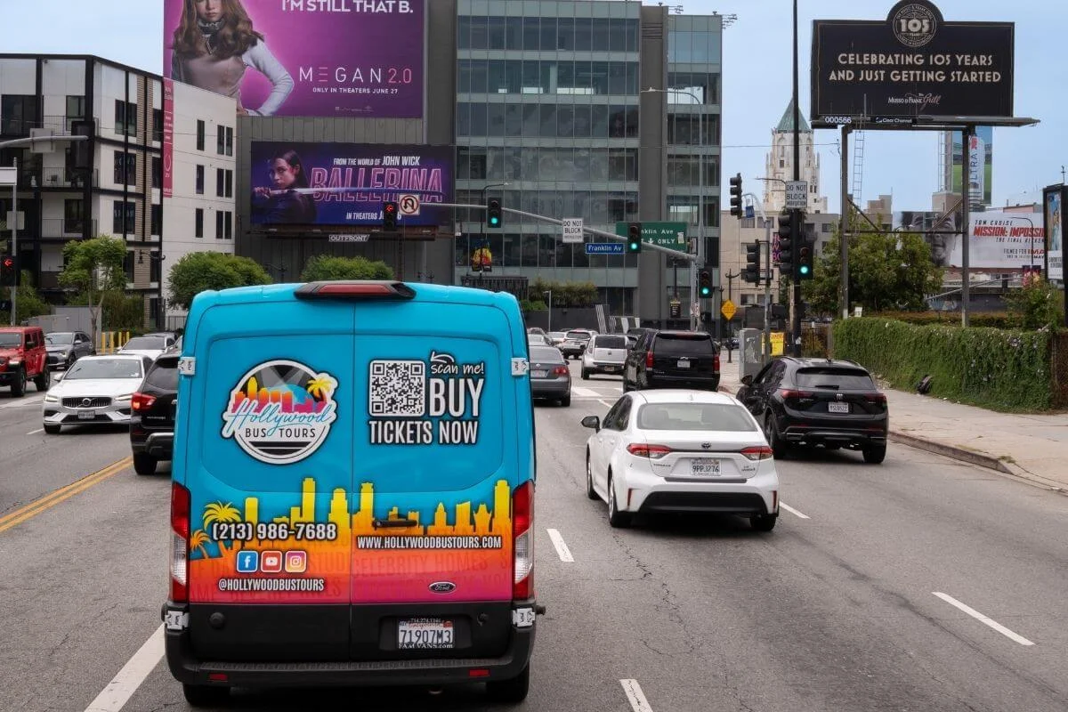 Bright blue Hollywood Bus Tours van driving down a busy Los Angeles street, surrounded by cars and tall buildings with large movie billboards in the background.