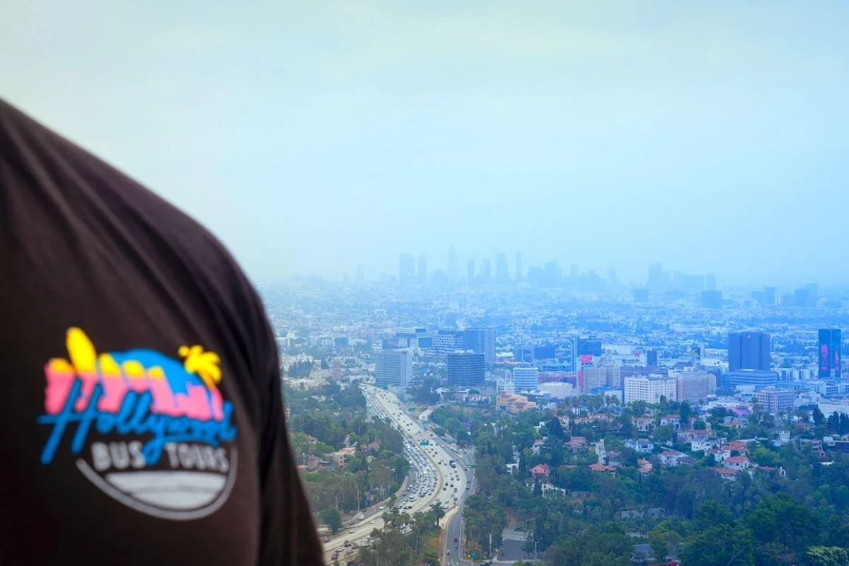 View from above of Los Angeles with a busy freeway leading toward the hazy downtown skyline, partially framed by the shoulder of a person wearing a “Hollywood Bus Tours” T-shirt in the foreground.