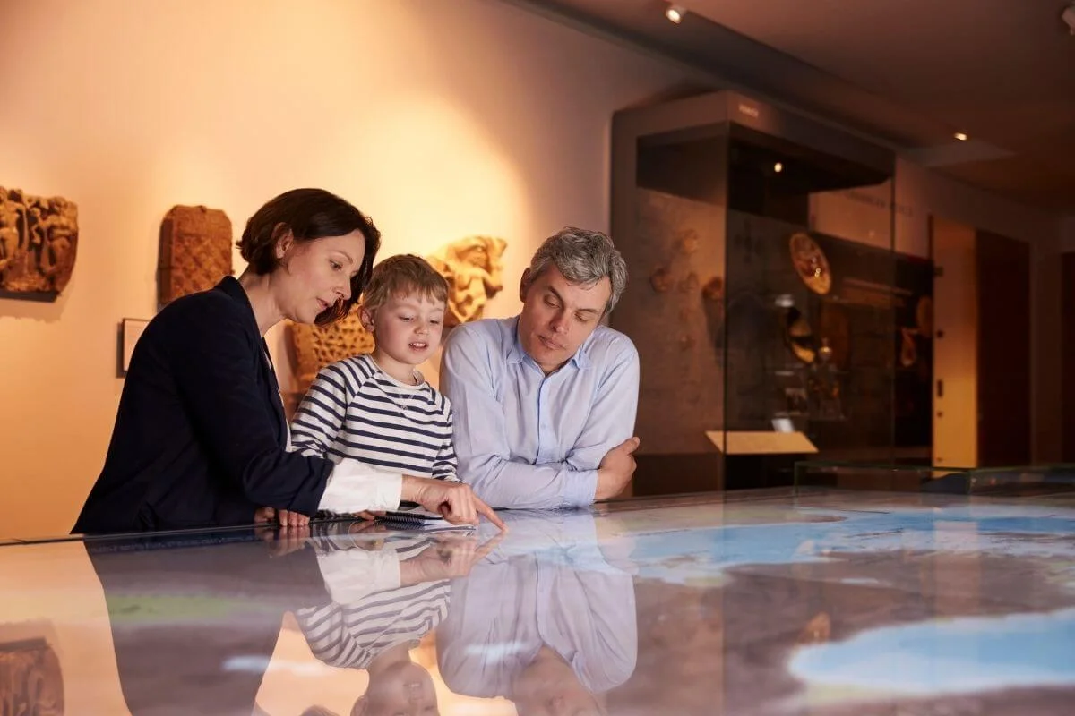 Mother, father, and young boy stand at a large interactive display table in a museum, with the mother pointing at a map while the child and father watch closely, surrounded by artifacts on the walls.