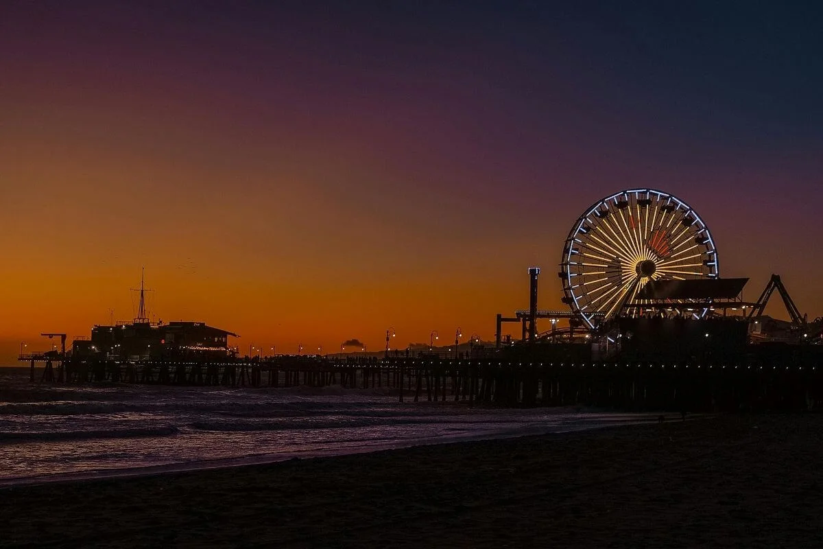 Silhouette of Santa Monica Pier with its illuminated Ferris wheel glowing against a vivid orange and purple sunset sky, with gentle waves rolling onto the darkened beach in the foreground.