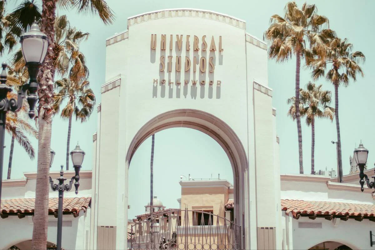 Daytime view of the grand white archway at the entrance to Universal Studios Hollywood, framed by tall palm trees and Spanish-style buildings under a pale blue sky.