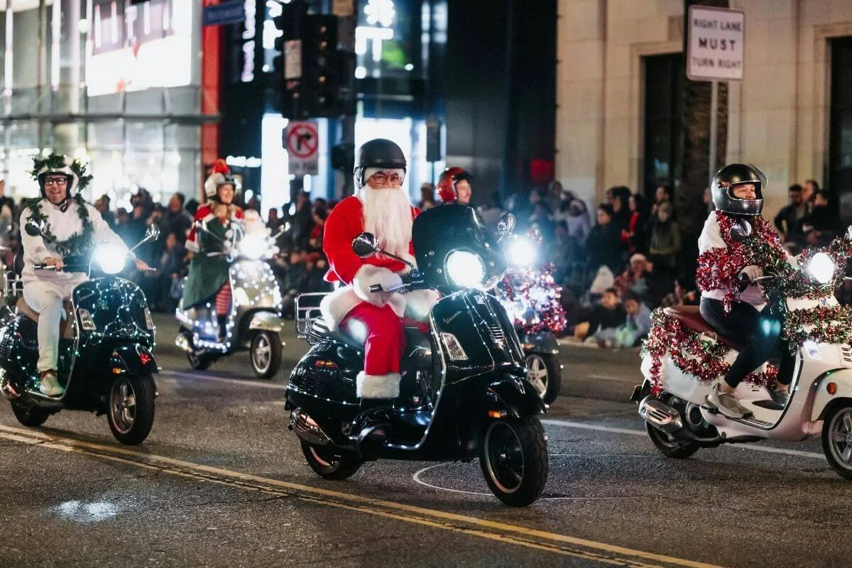 Nighttime street parade featuring people in festive costumes, including someone dressed as Santa Claus, riding decorated scooters with bright headlights and tinsel, while crowds watch from the sidewalks.