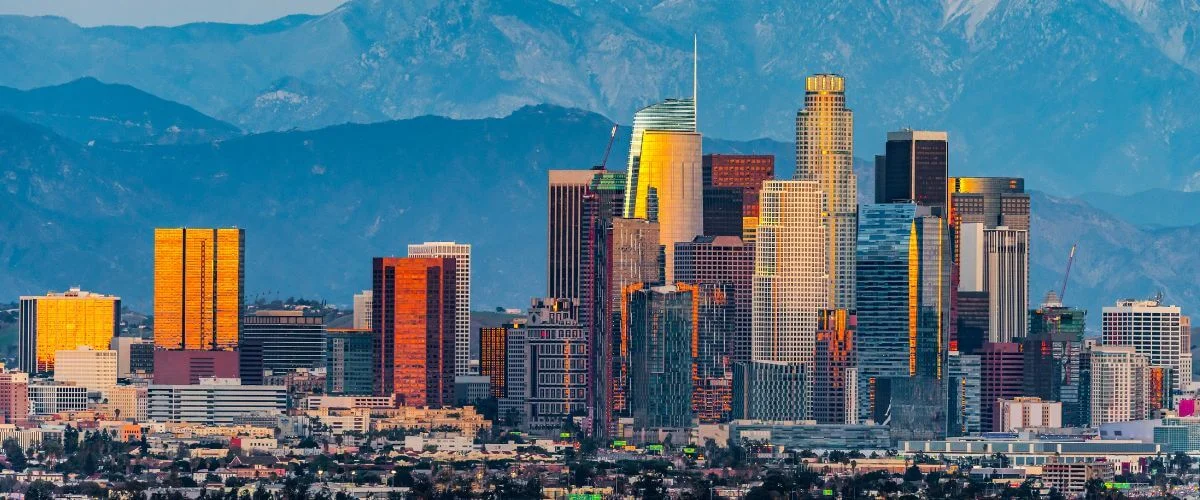 Panoramic view of downtown Los Angeles high rise buildings glowing in warm sunset light with hazy blue mountains in the background.