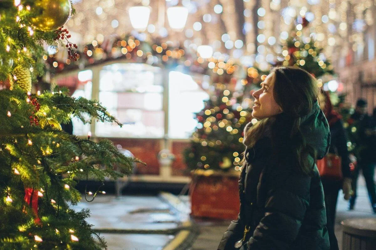 Young woman in a puffy winter coat smiles while looking at a decorated Christmas tree covered in lights and ornaments at a festive outdoor market, with blurred holiday lights glowing in the background.