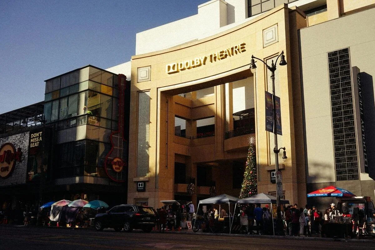 Daytime street view of the Dolby Theatre entrance in Hollywood, with a decorated Christmas tree, vendor tents, and people walking along the sidewalk, next to the neighboring Hard Rock Cafe building.
